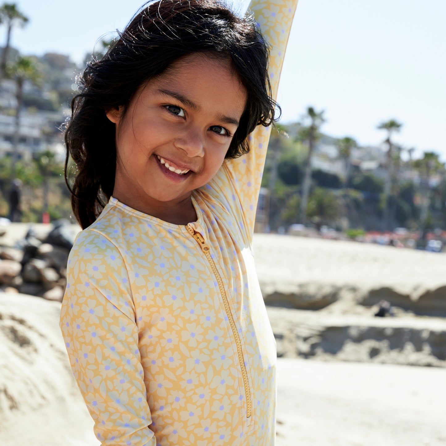 Young girl in a yellow florish patterned swimsuit with long sleeves and a zip standing on a beach with palm trees in the background