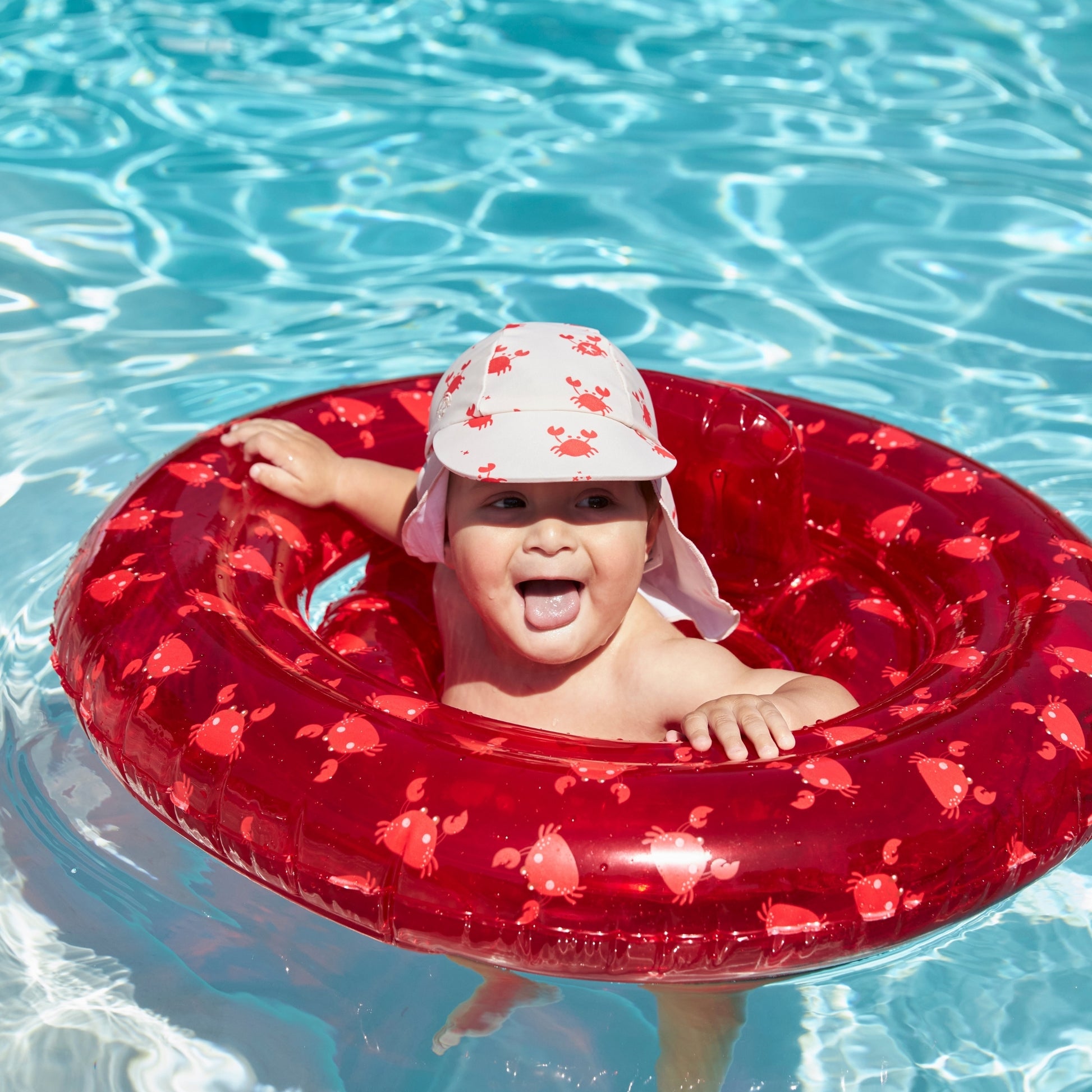 Child sticking out his tongue in the swimming pool in a red crabby inflatable baby float with a crabby sun cap with a protective flap that shields the skin