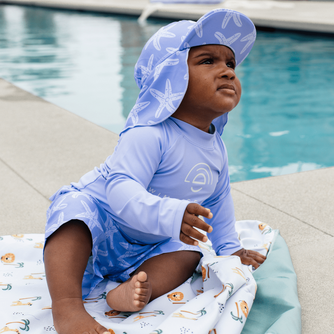 Child sitting on the ground at the pool wearing a lila swimming T-shirt and a Lila Seastar swim short with a Lila Seastar Sun cap with a flap that shields the skin.