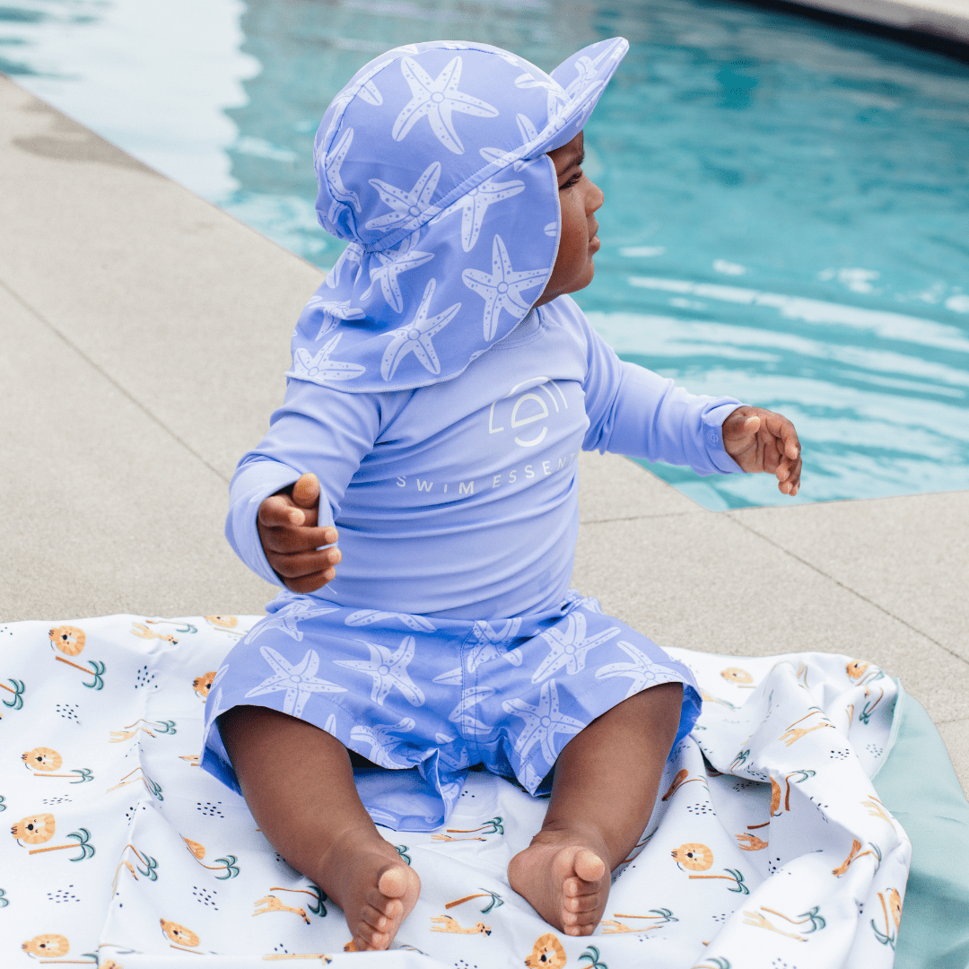 Child sitting on the ground by the pool, looking to the right, wearing a lila swim T-shirt, Lila Seastar swim shorts, and a Lila Seastar sun cap with a protective neck flap.