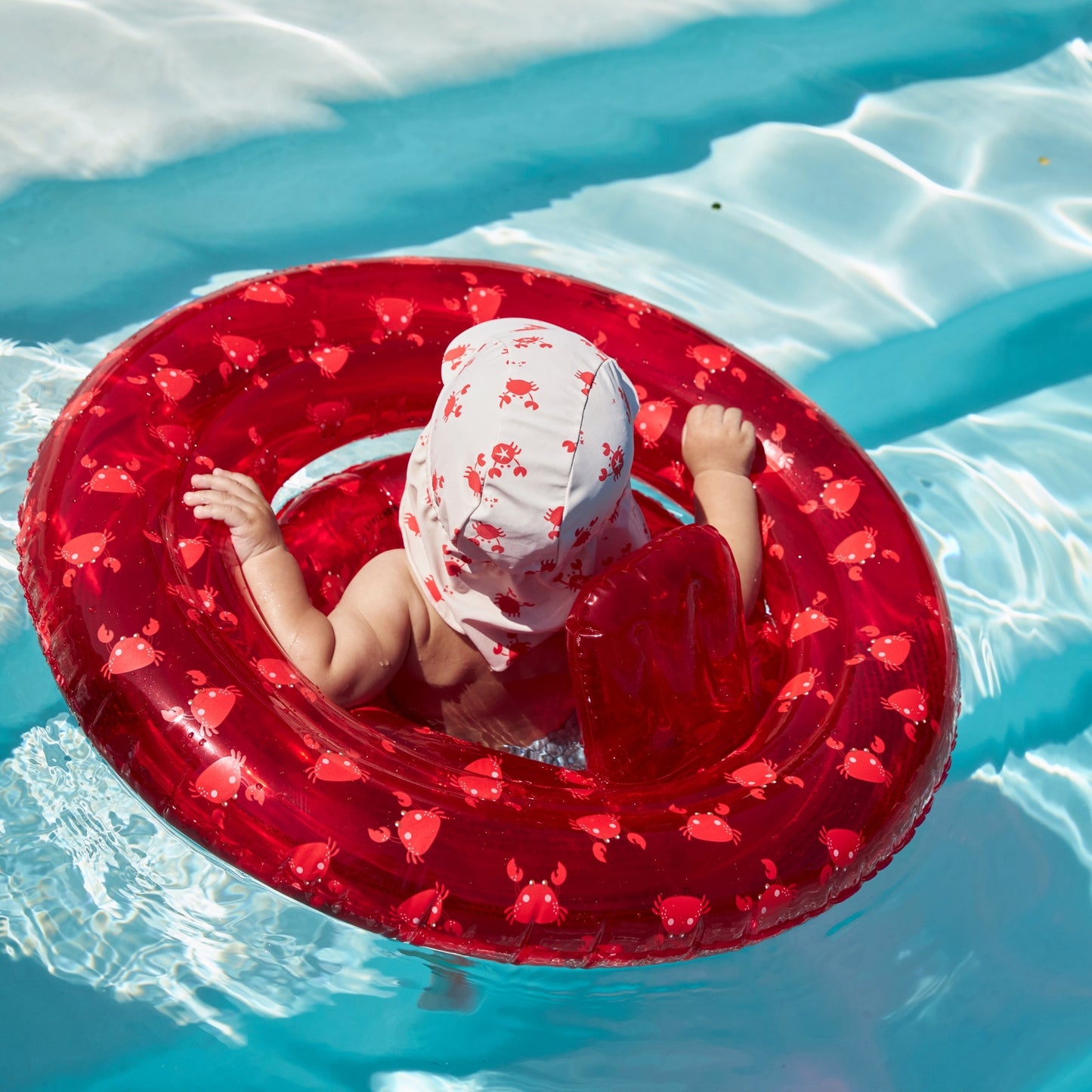Child from the back in the swimming pool in a red crabby inflatable baby float with a sun hat with a protective flap that protects the skin.