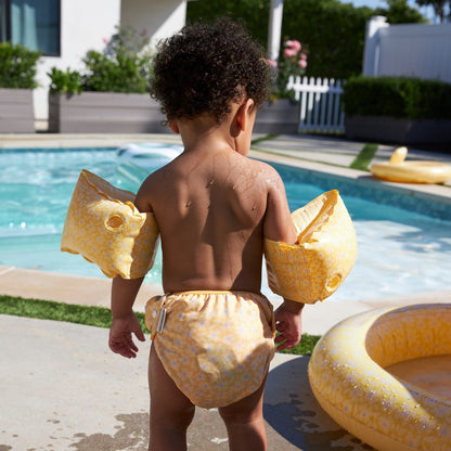 Child with yellow florish armbands and yellow florish swim diaper standing by a pool
