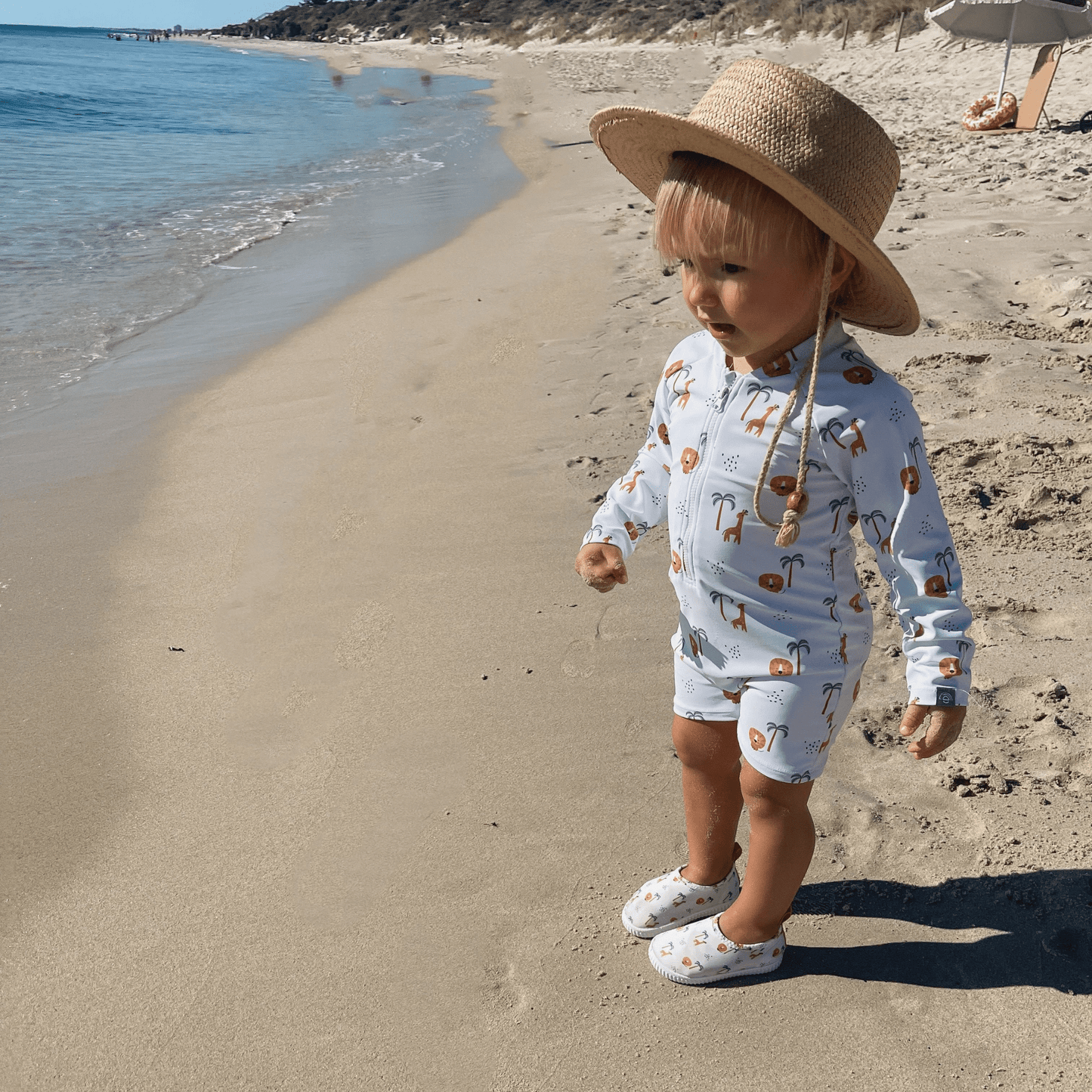 A child wearing a long-sleeve jungle- and animal-print swimsuit, matching water shoes, and a beige sun hat, standing on a sandy beach.