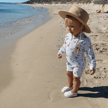 A child wearing a long-sleeve jungle- and animal-print swimsuit, matching water shoes, and a beige sun hat, standing on a sandy beach.