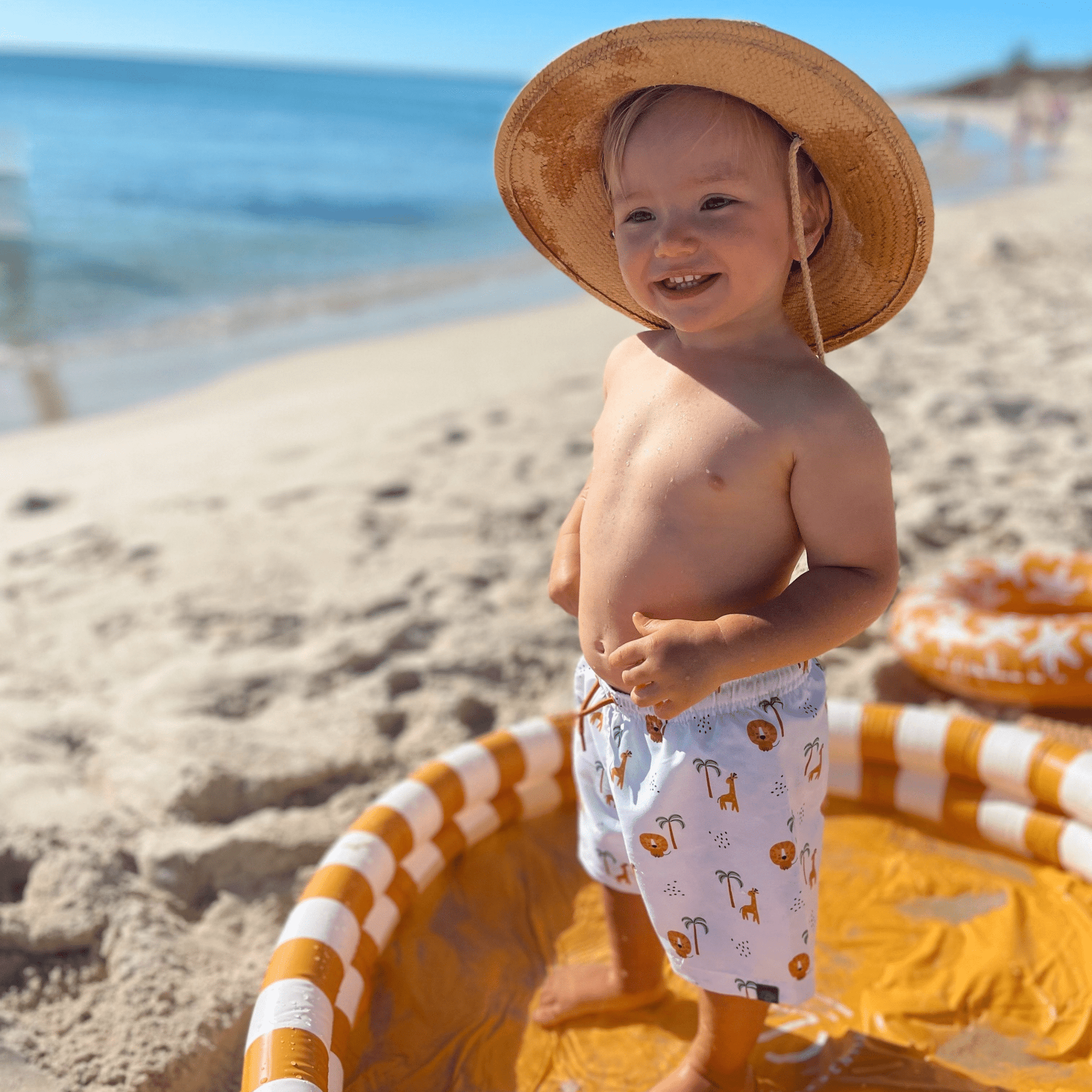Child on a beach wearing a straw hat and swim shorts with a pool float in the foreground.