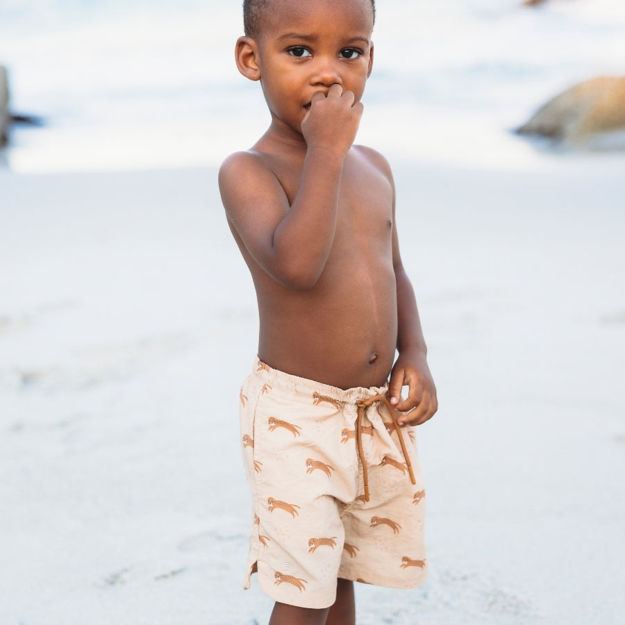 Young child wearing brown swimming shorts with brown leopard prints on a beach.