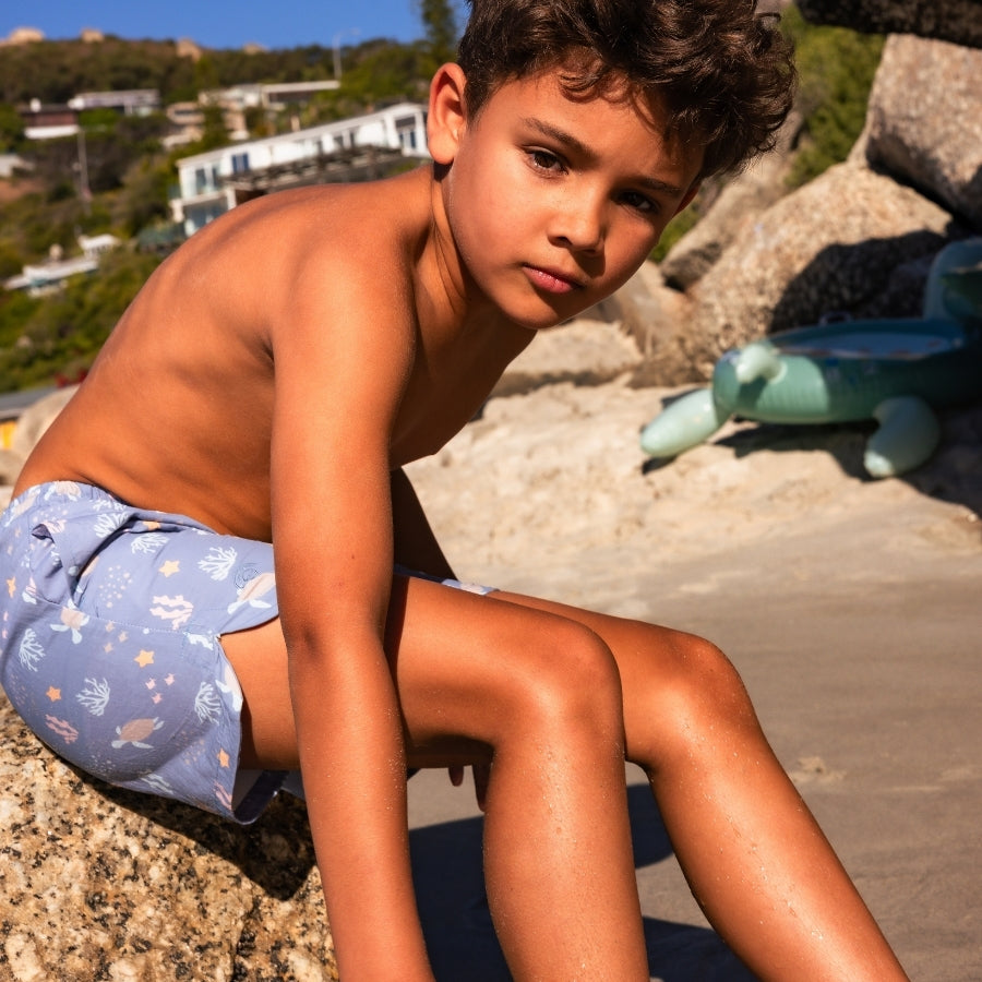 Young boy sitting on a rock by the beach with a toy dinosaur.