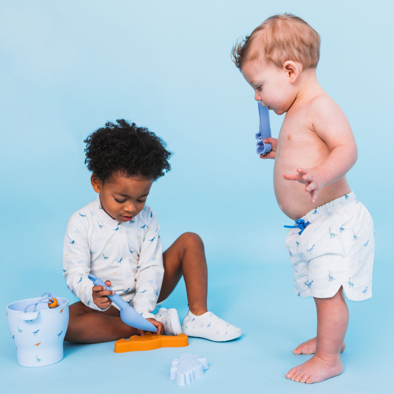 Two children playing with toys wearing the dino swimshirt and swim short on a blue background