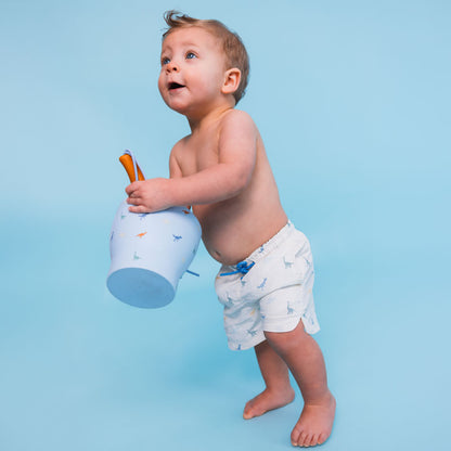 Baby holding a toy bucket against a blue background wearing a dino swimshort