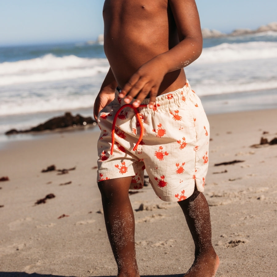 A child wearing beige swim shorts with a crab print on a beach with ocean waves in the background