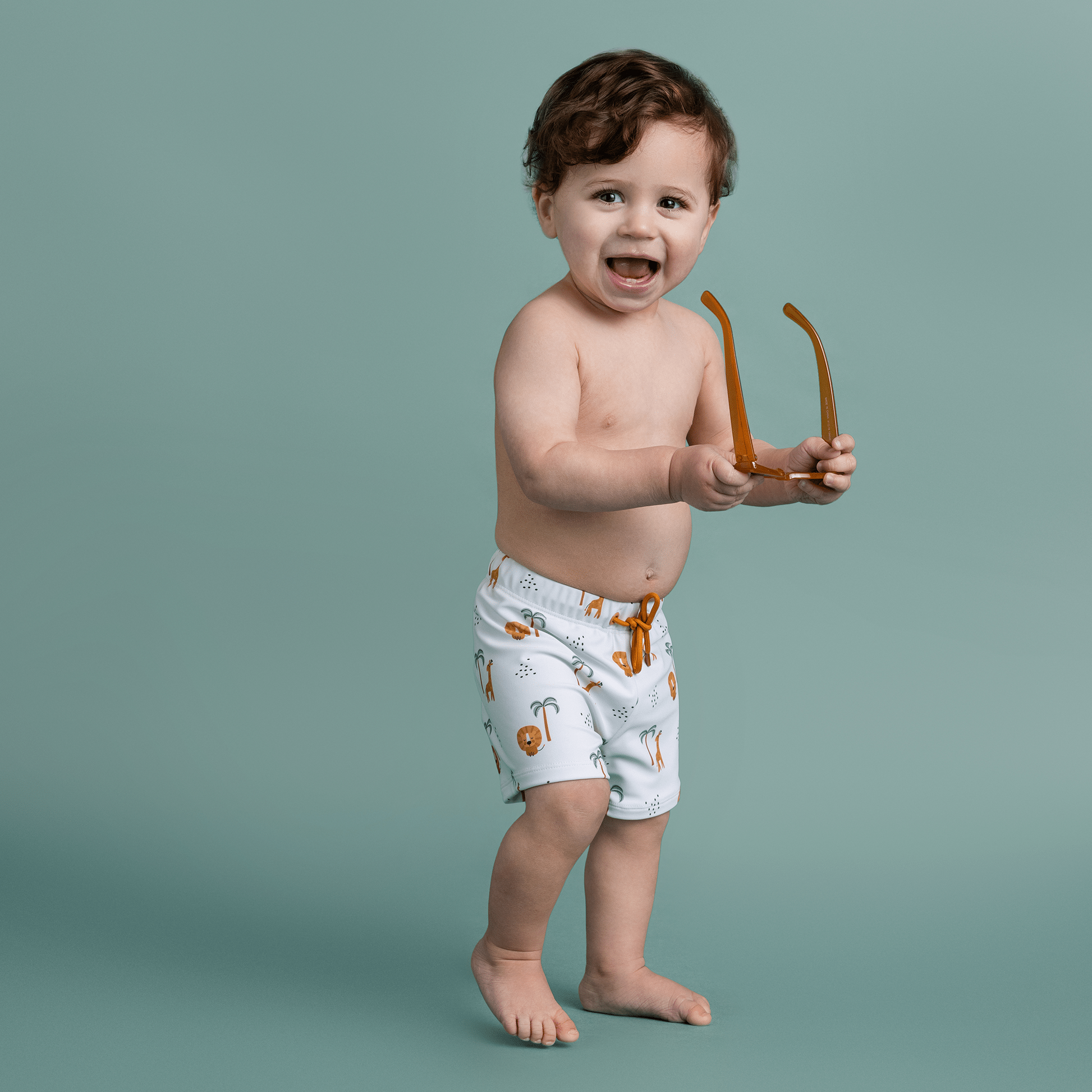 Child wearing patterned swim shorts holding sunglasses on a green background
