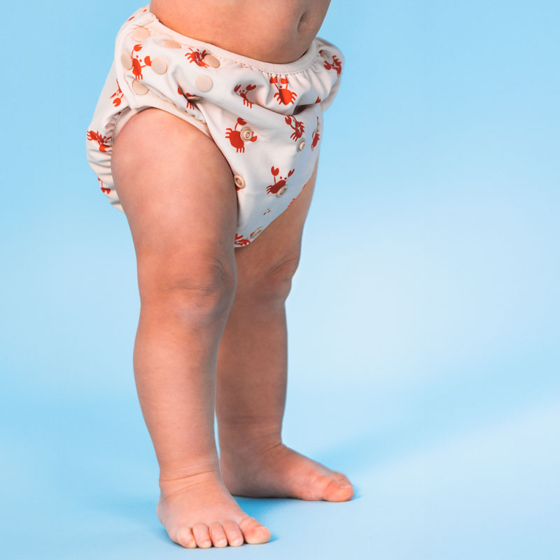 Baby wearing beige swim diaper with red crab pattern on a blue background