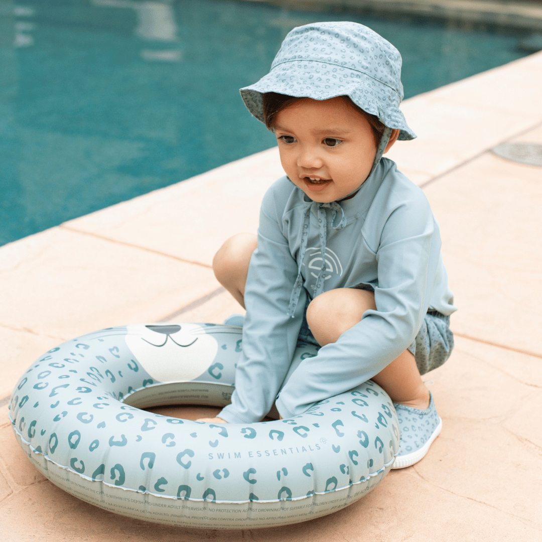 Child wearing a light blue outfit and hat, sitting next to an inflatable pool ring by a pool.