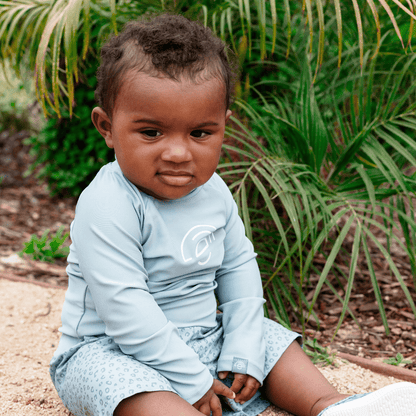 Child wearing a light blue long-sleeve swimming shirt and shorts sitting outdoors with greenery in the background