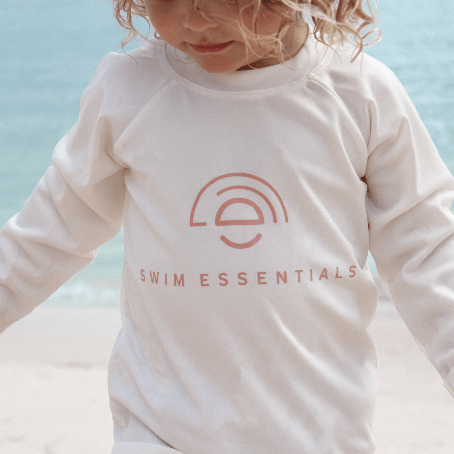 Child wearing a white long-sleeve shirt with 'Swim Essentials' logo against a light blue background