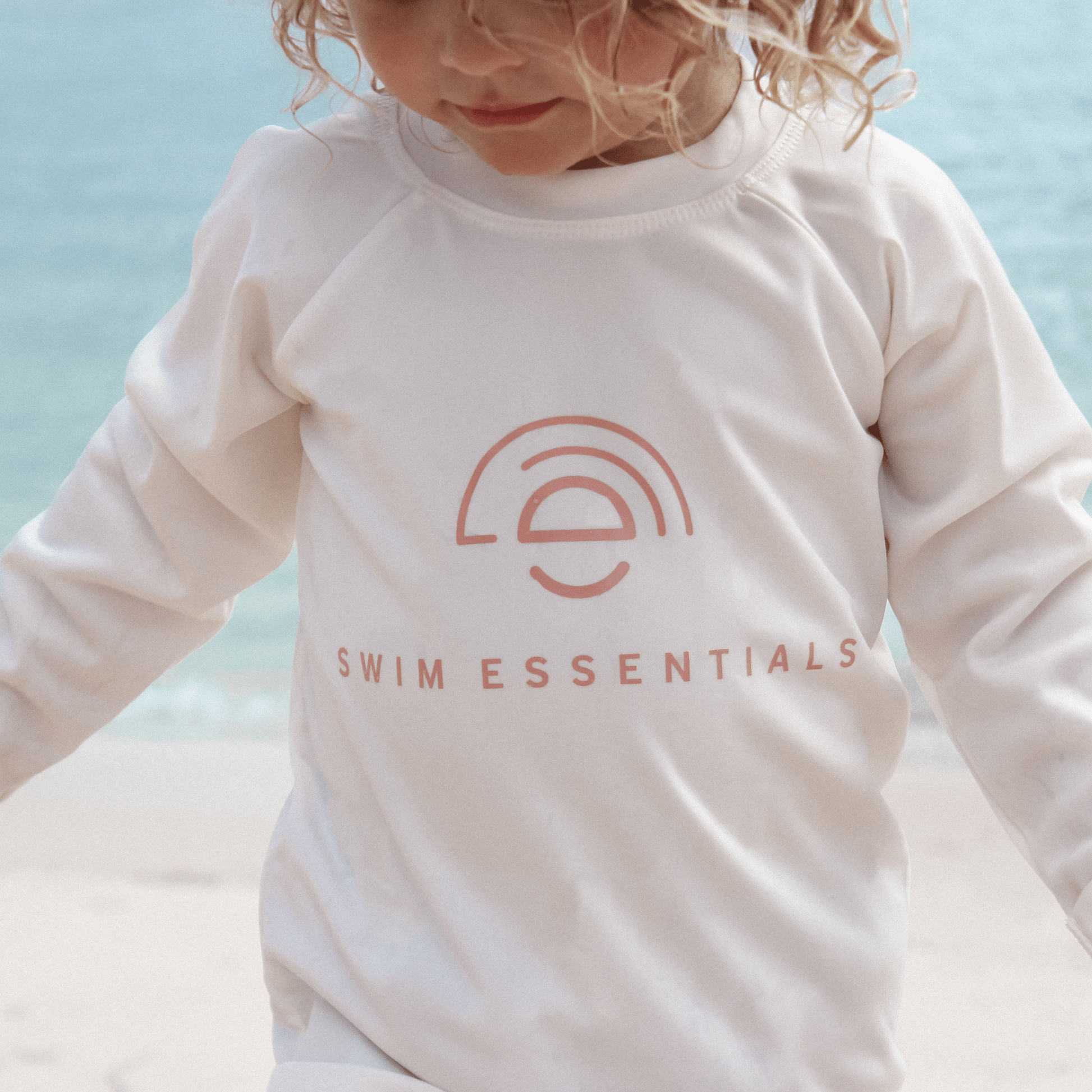Child wearing a white long-sleeve shirt with 'Swim Essentials' logo against a light blue background