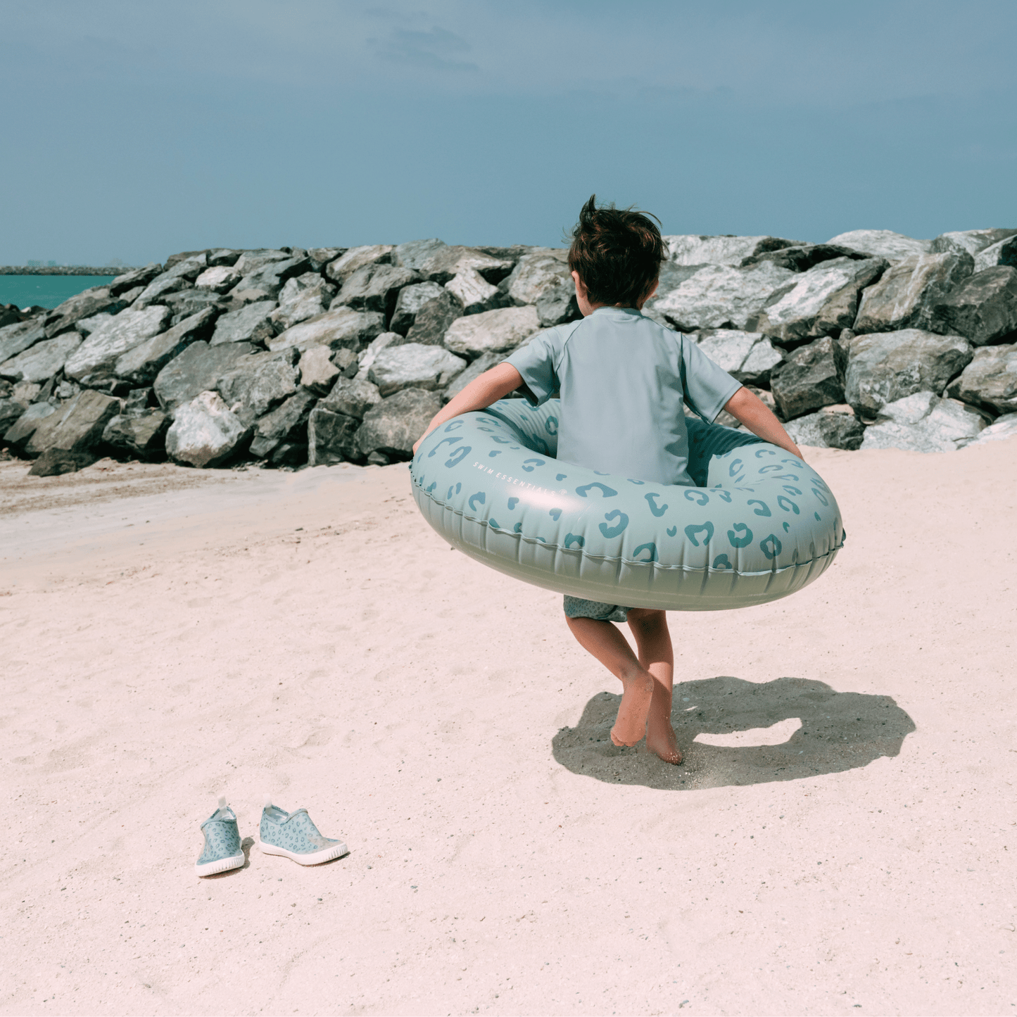 Child holding a blue inflatable ring on a sandy beach with rocks and water in the background