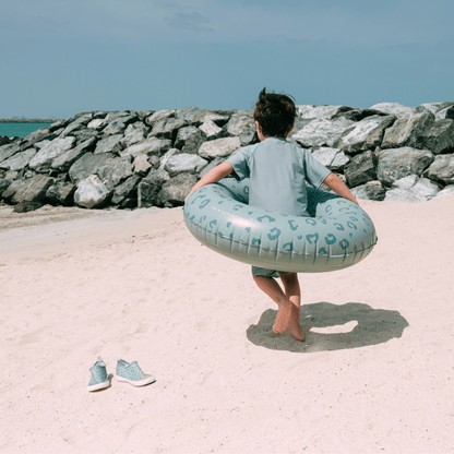 Child holding a blue inflatable ring on a sandy beach with rocks and water in the background