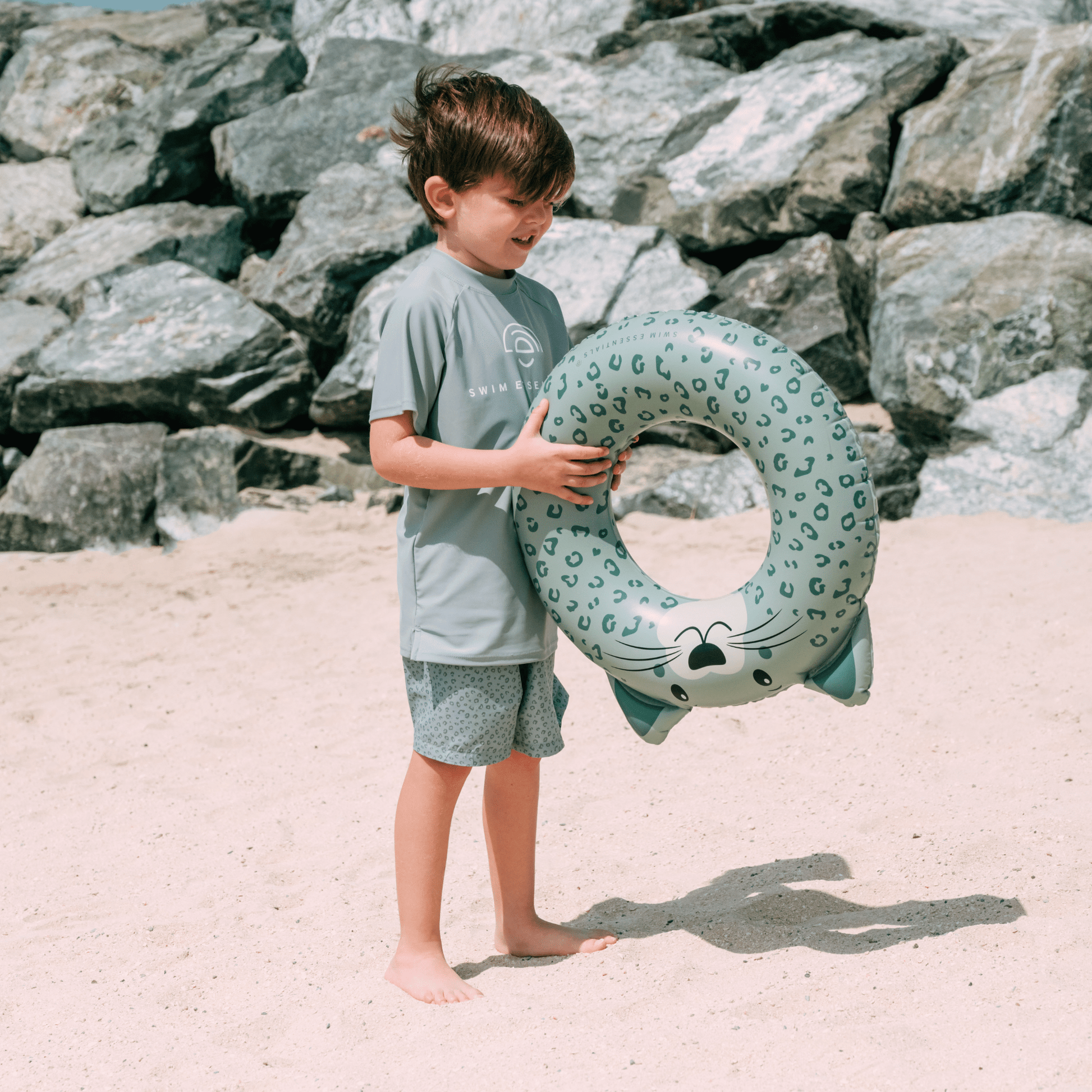 Child holding a shark-shaped inflatable ring on a beach with rocks in the background