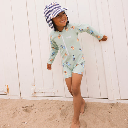 Child wearing a light green swimsuit with cartoon dog design and a striped white-blue hat on a sandy beach.