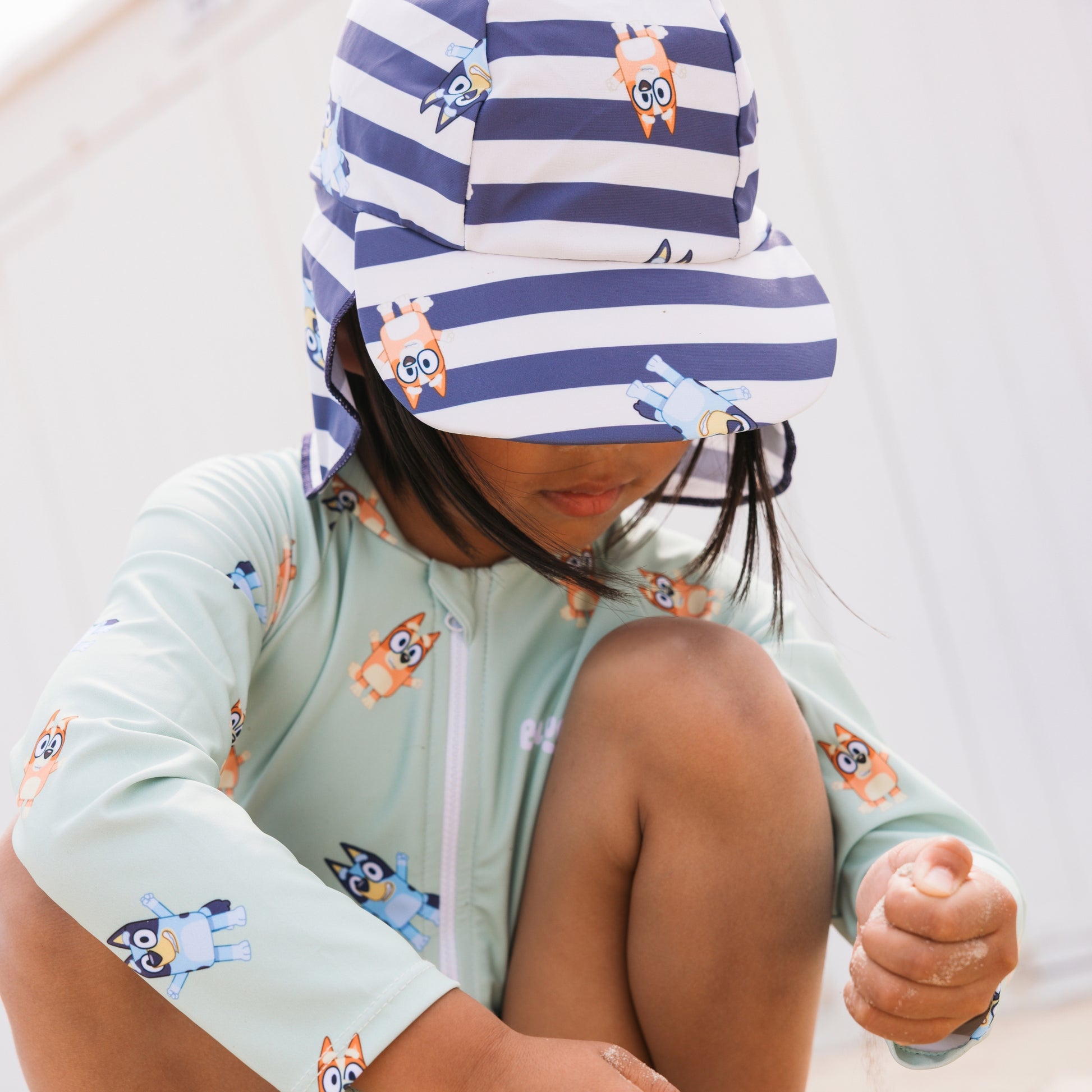 Child wearing a light green bluey swimsuit long-sleeve with cartoon dog design and a striped hat blue-white with animal prints.