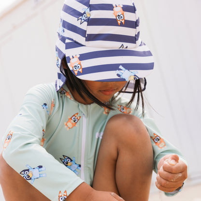 Child wearing a light green bluey swimsuit long-sleeve with cartoon dog design and a striped hat blue-white with animal prints.