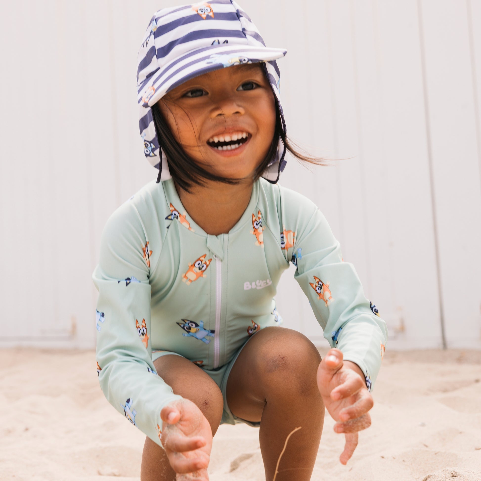 Child wearing a Bluey light green long-sleeve shirt and striped blue-white hat, sitting on a sandy surface.