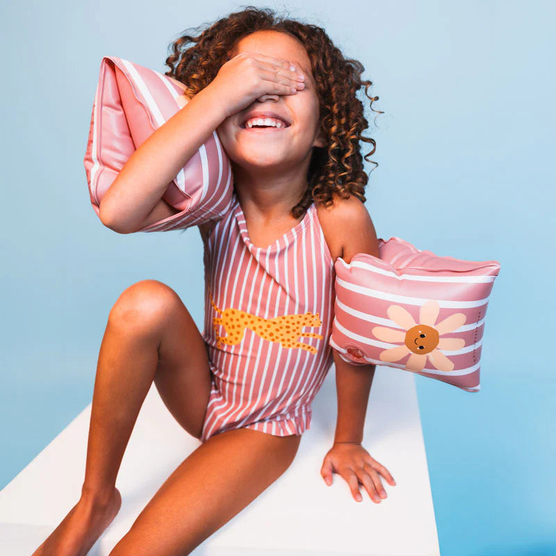 Child wearing a pink and white striped swimsuit with floral arm bands against a light blue background