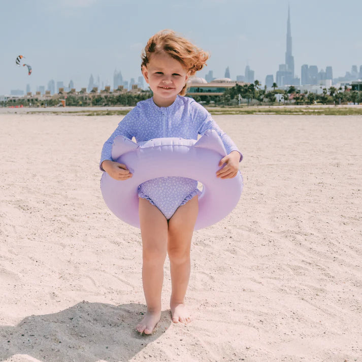Child wearing a leopard swimsuit holding a purple inflatable ring on a sandy beach with a city skyline in the background
