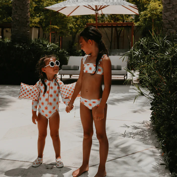 Two young girls in matching swimsuits with the floral pattern standing on a garden with outdoor furniture and plants in the background.