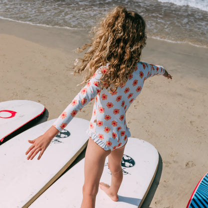 Child in a floral swimsuit standing on a beach with surfboards.
